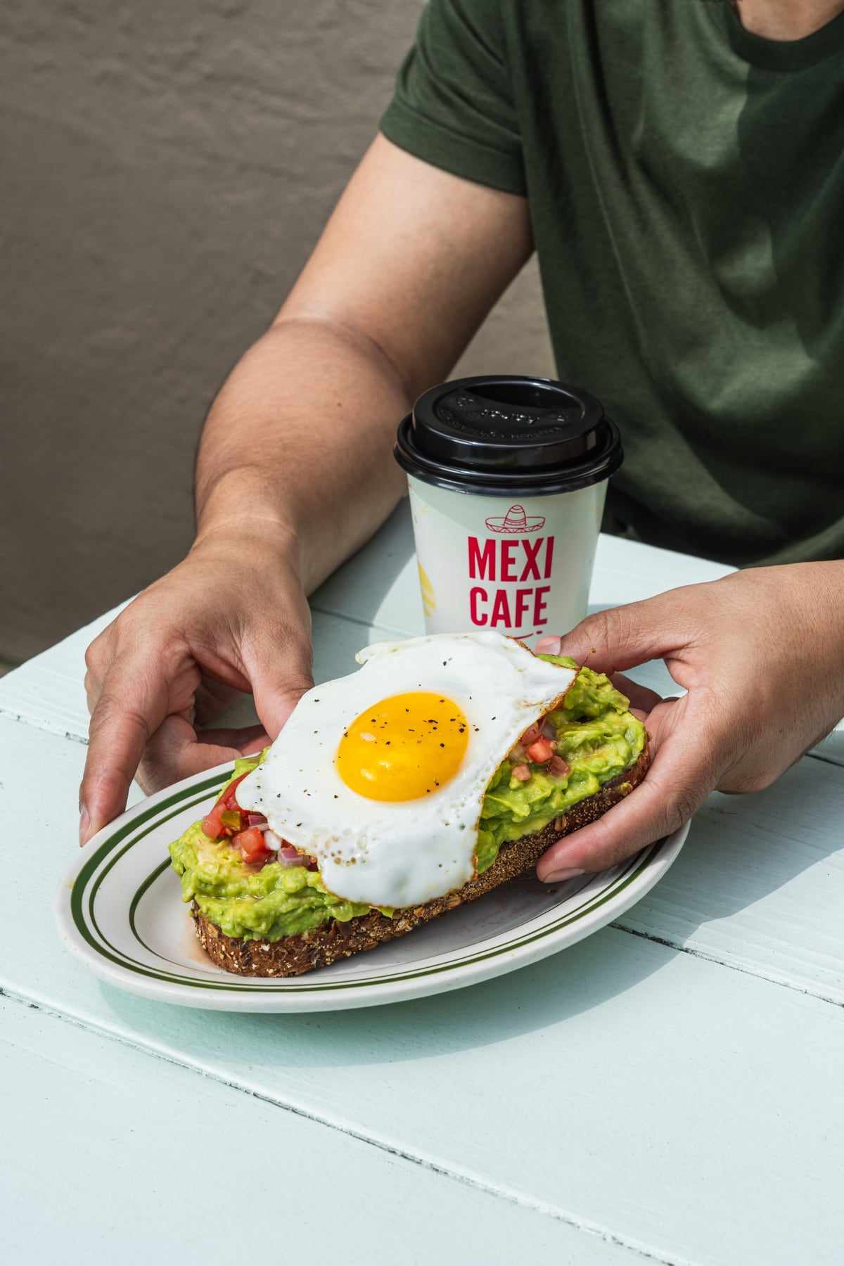 Avocado toast with a fried egg and pico de gallo held over a plate, next to a Mexi Cafe coffee cup