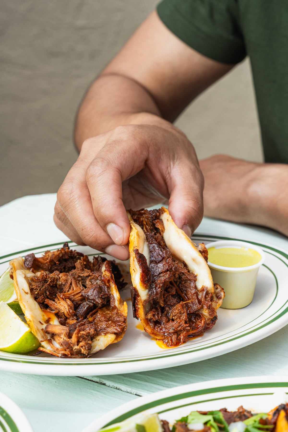 Two birria tacos held over a plate, the beef glistening, with a cup of green salsa