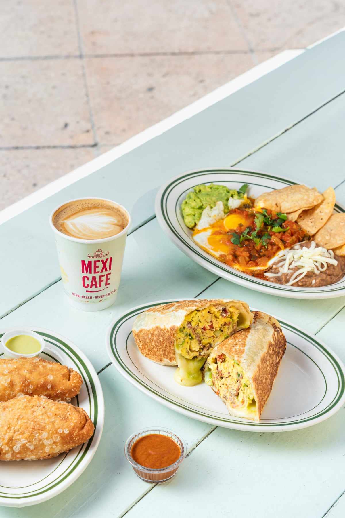 Breakfast at the window: a cortadito in a Mexi Cafe cup, huevos rancheros and a burrito on a pale blue table