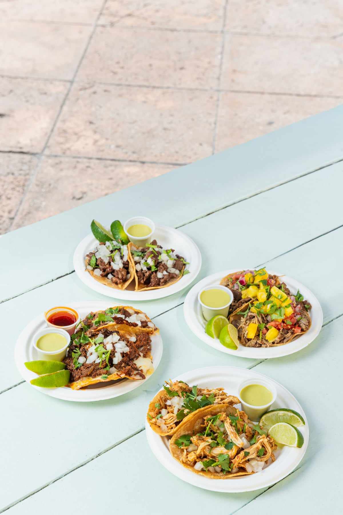 Three plates of street tacos with salsas and lime on a pale blue table